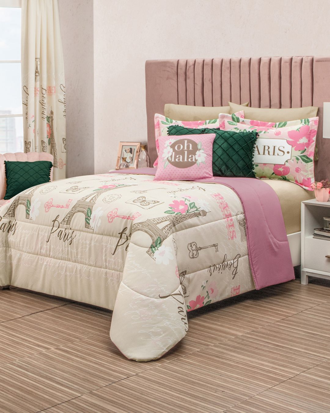 A neatly made bed features the Juego de Edredón Súper Suave Ciudad Luz by Intima Hogar, with Paris-inspired design and pillows in cream, pink, and green. The upholstered headboard and lamp enhance the room’s bright, cozy feel.