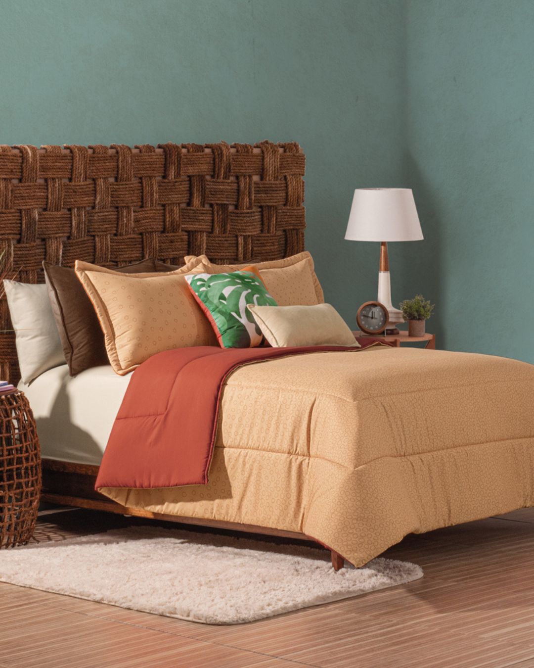 A cozy bedroom featuring the Intima Hogar Juego de Edredón Súper Suave Fresno, a woven wicker headboard, decorative pillows, a bedside table with a lamp, books, clock, and plant set against a teal wall.