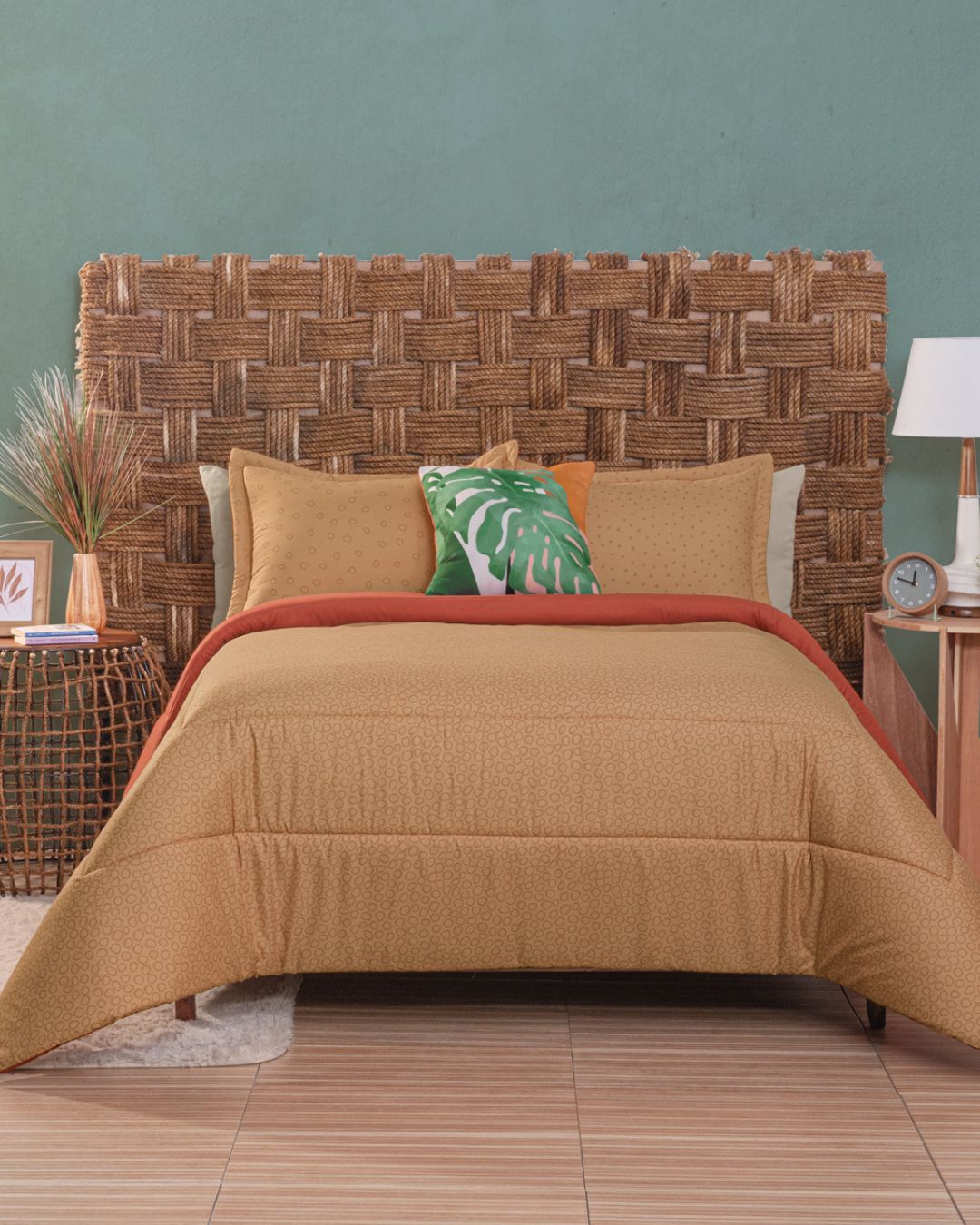 A bed with a woven wicker headboard, Juego de Edredón Súper Suave Fresno by Intima Hogar in tan, an orange throw, and green leaf print decorative pillow. Next to it: a round wicker side table with plants and a wooden nightstand with lamp and clock.