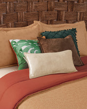 A bed styled with the Juego de Edredón Súper Suave Fresno by Intima Hogar displays tan and rust bedding, five decorative pillows in green, brown, cream, and a leaf pattern, all set against a woven headboard.