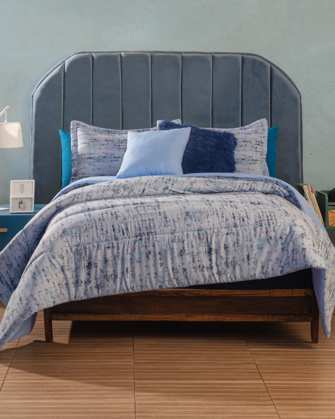 A bed with a tall, curved, upholstered blue velvet headboard features the Juego de Edredón Súper Suave Praga by Intima Hogar in blue and white, several decorative pillows, and sits on a wooden frame against a light blue wall and wooden floor.