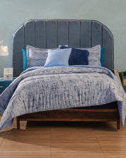 A bed with a tall, curved, upholstered blue velvet headboard features the Juego de Edredón Súper Suave Praga by Intima Hogar in blue and white, several decorative pillows, and sits on a wooden frame against a light blue wall and wooden floor.