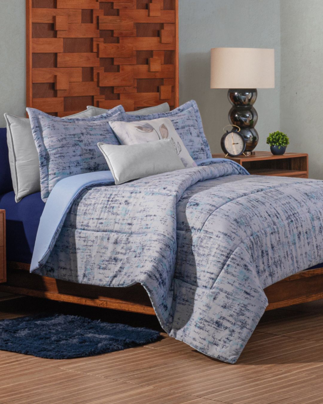 A neatly made bed features the Juego de Edredón Súper Suave Praga by Intima Hogar in blue and white, with decorative pillows, a blue rug, wooden headboard, nightstand with lamp, clock, and small plant in the background.