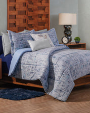 A neatly made bed features the Juego de Edredón Súper Suave Praga by Intima Hogar in blue and white, with decorative pillows, a blue rug, wooden headboard, nightstand with lamp, clock, and small plant in the background.