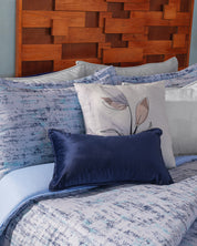A neatly made bed features the Juego de Edredón Súper Suave Praga by Intima Hogar in blue and white, a blue velvet lumbar pillow, decorative pillows with leaf designs, and a wooden headboard with a geometric pattern.