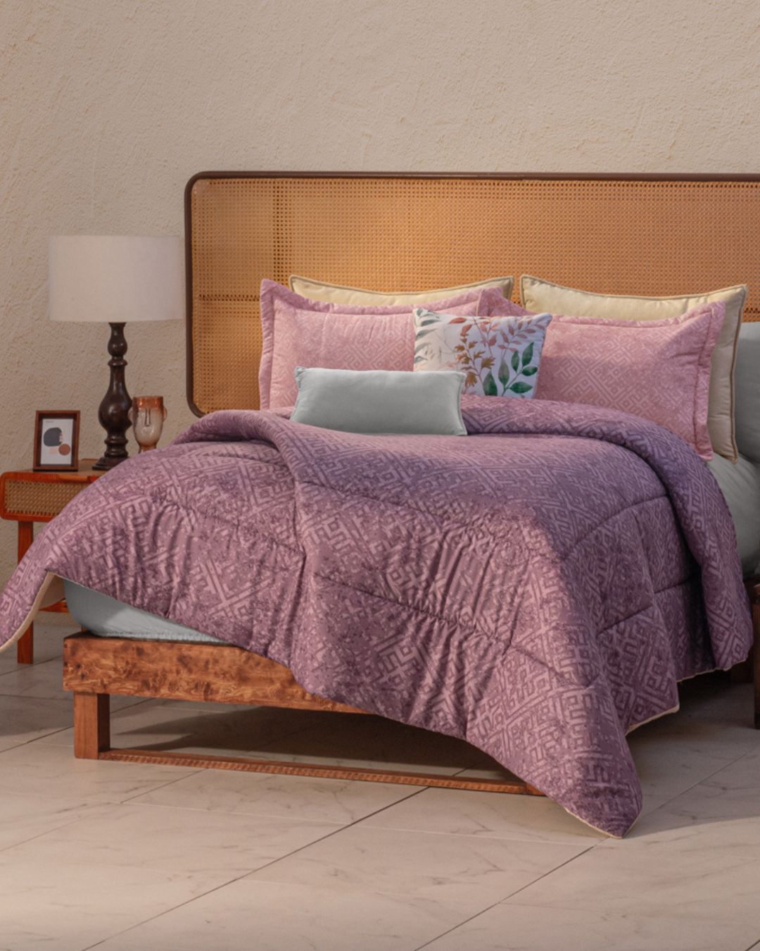 A neatly made bed with a wooden frame and rattan headboard features the Juego de Edredón Súper Suave Toronto by Intima Hogar in mauve, pastel decorative pillows, a blue lumbar pillow, and a nearby nightstand with lamp and photo—ideal for decoración contemporánea.