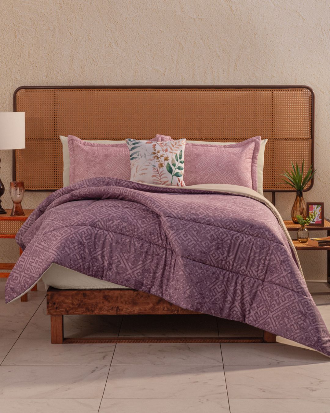 A neatly made bed with a light brown wicker headboard, mauve pillows, the Juego de Edredón Súper Suave Toronto by Intima Hogar, and wooden side tables with lamps and plants create una decoración contemporánea against the beige wall.