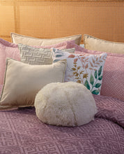 A neatly made bed features the Juego de Edredón Súper Suave Toronto by Intima Hogar, in mauve with matching pillows and decorative cushions—including a white furry round pillow and a floral one—set against a woven rattan headboard.