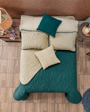 A neatly made bed with the Juego de Edredón Velvet Vida by Intima Hogar, featuring green and beige quilted bedding, two matching beige pillows, a green accent pillow, and a woven beige pillow beside a wooden nightstand.
