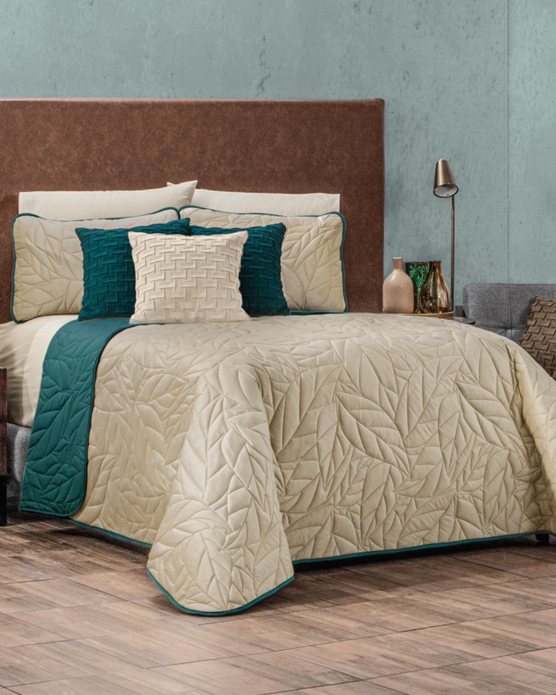 A neatly made bed features the Intima Hogar Juego de Edredón Velvet Vida, a beige leaf-patterned reversible set with teal accents and matching fundas decorativas. A brown headboard and modern nightstand decor complete the stylish look.
