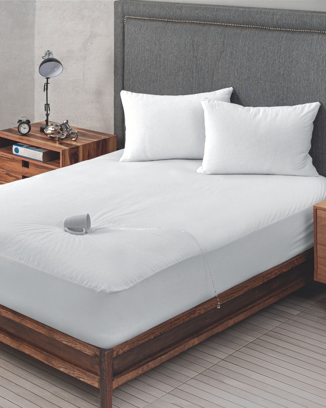 A neatly made bed with white bedding features a small water spill on the mattress, but thanks to the Protector de Colchón Impermeable by Intima Hogar, the surface stays clean and perfect for un descanso fresco.