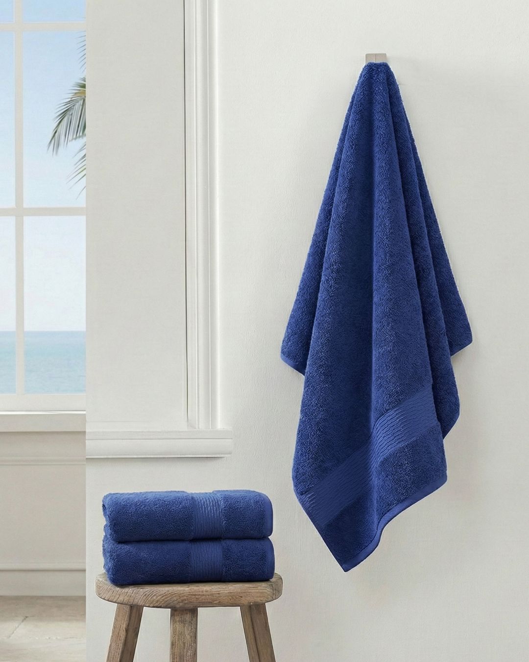 A Toalla de Baño Americana Azul de Intima Hogar cuelga en una pared blanca junto a una ventana con vista al mar; dos más, reconocidas por su absorbente comodidad, están dobladas sobre un banco de madera abajo.