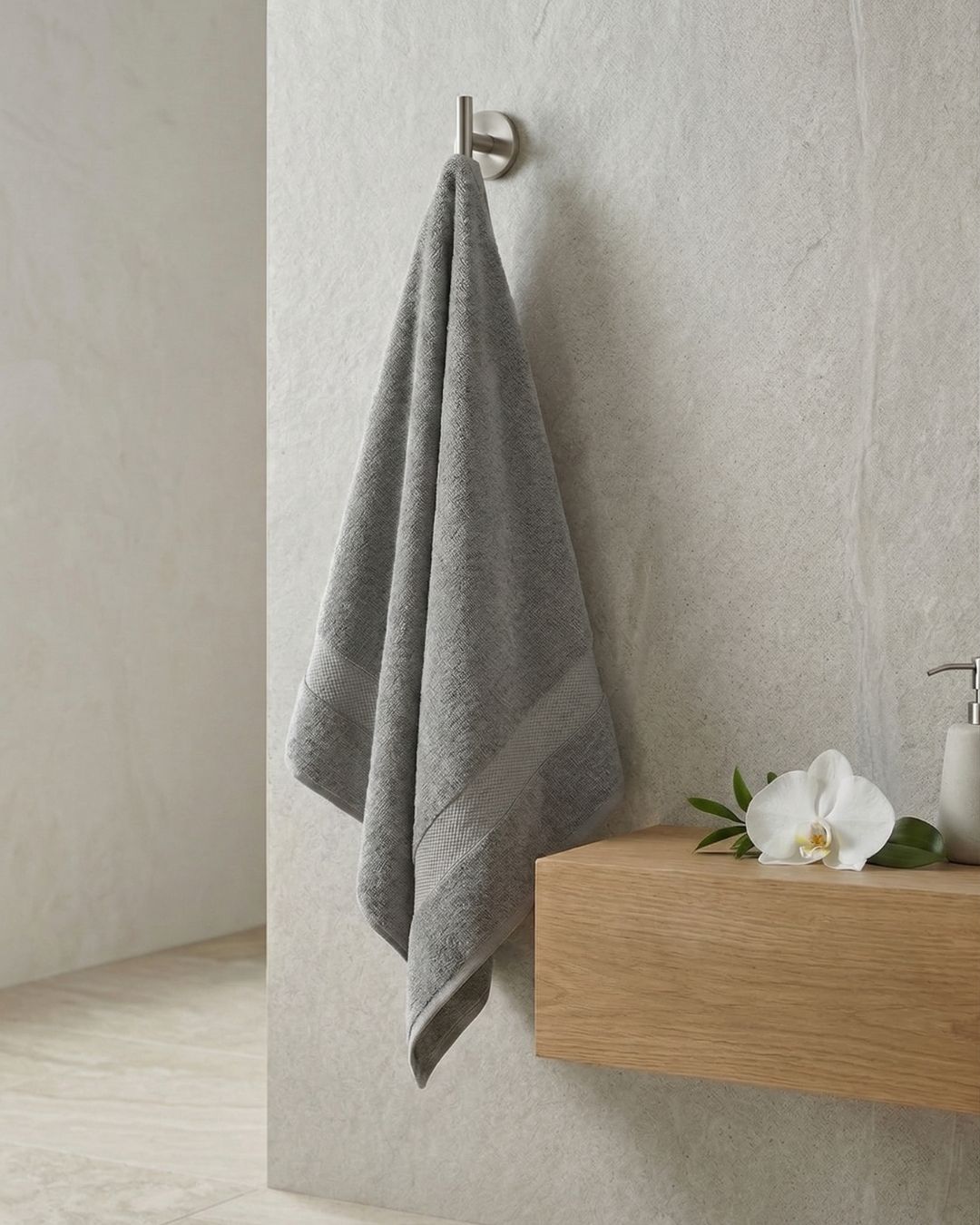La Toalla de Baño Americana Gris de Intima Hogar cuelga de un gancho en una pared clara, cerca de una superficie de madera con orquídea blanca, hojas verdes y un dispensador, creando un ambiente acogedor.