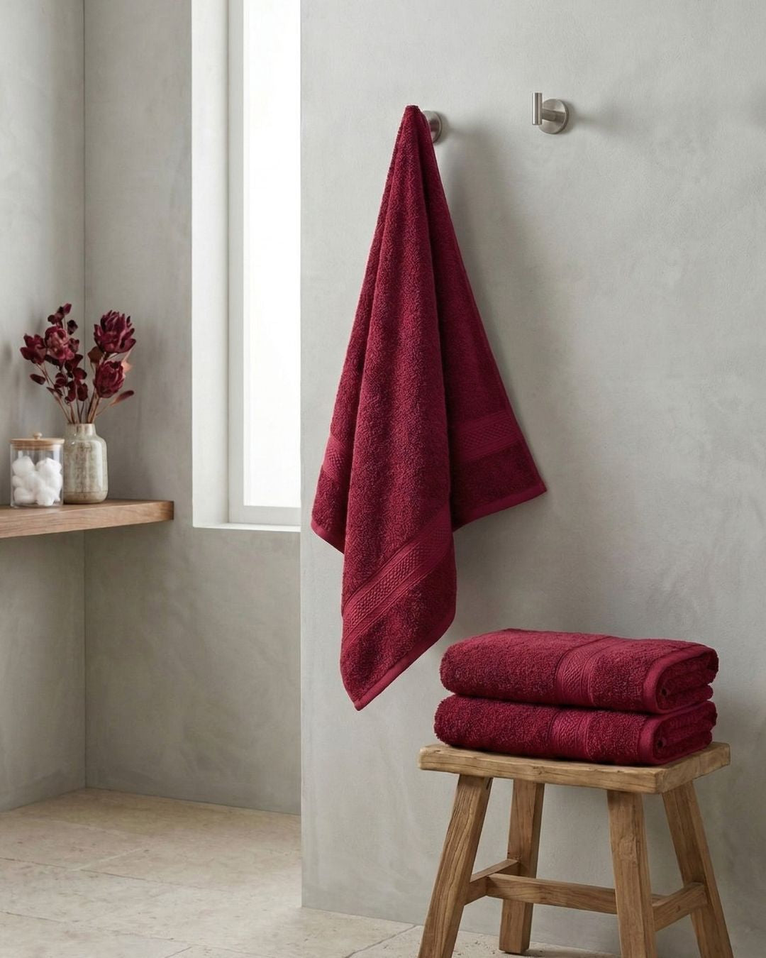 La Toalla de Baño Americana Vino de Intima Hogar cuelga en un baño minimalista con muros gris claro, sobre un banco de madera con toallas a juego, cerca de una ventana y una repisa con flores rojas y un frasco de algodones.