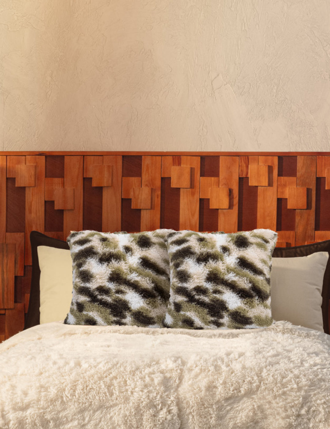 A cozy bed with Intima Hogar’s Juego de Cojines de Lujo Finlandia—one with a spotted print, another long-pile—paired with a wooden geometric headboard, all set against a beige textured wall.