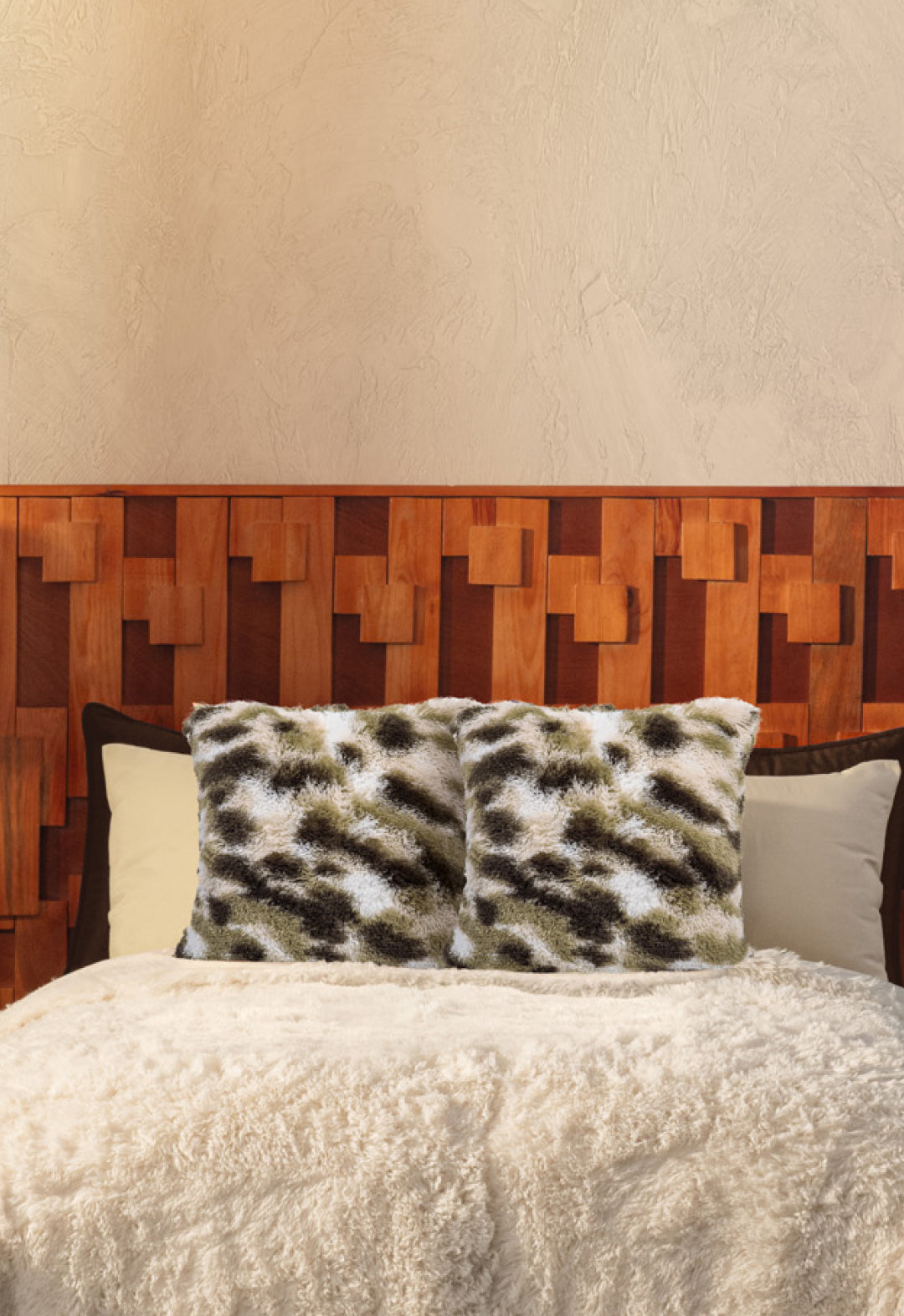 A cozy bed with Intima Hogar’s Juego de Cojines de Lujo Finlandia—one with a spotted print, another long-pile—paired with a wooden geometric headboard, all set against a beige textured wall.