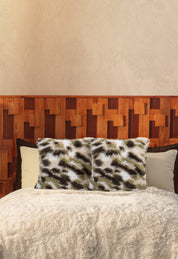 A cozy bed with Intima Hogar’s Juego de Cojines de Lujo Finlandia—one with a spotted print, another long-pile—paired with a wooden geometric headboard, all set against a beige textured wall.