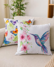 Two decorative pillows on a beige sofa: one is the Cojin Mariposa by Intima Hogar with colorful butterflies and leaves, the other features a vibrant hummingbird and pink flowers. Both are watercolor style, made from microfibra with synthetic fiber filling.
