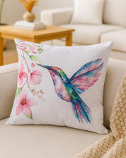 The Cojin Mariposa by Intima Hogar is a white throw pillow with a vibrant watercolor hummingbird and pink flowers, filled with suave fibra sintética, displayed on a beige sofa in a bright, cozy living room.