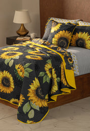 A neatly made bed with the Coordinado de Edredón Ultra Suave Amarelo by Intima Hogar, featuring bright yellow sunflowers and green leaves, sits beside a wooden nightstand with a lamp, books, and a cup—perfect for your room.