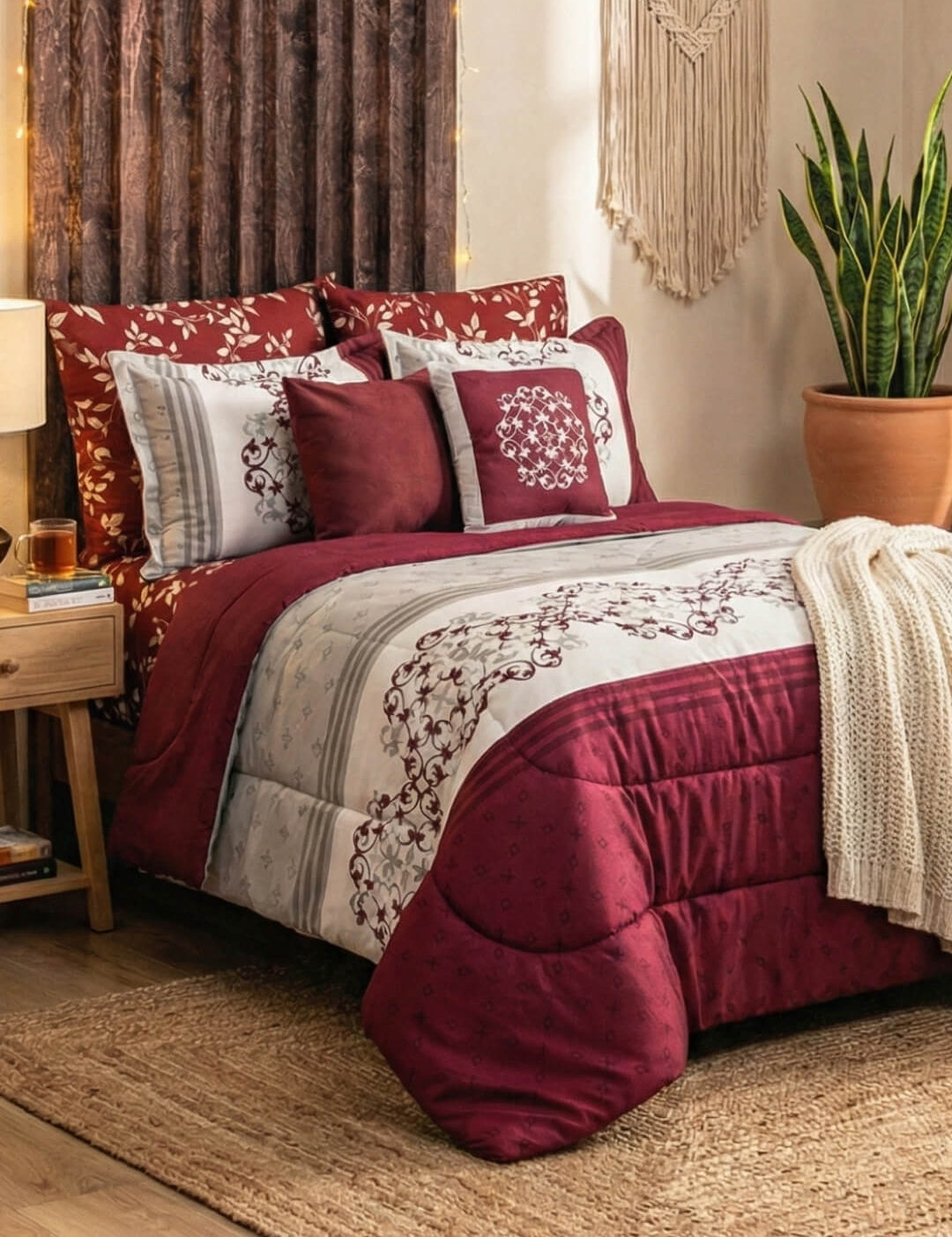 A cozy bedroom with the Intima Hogar Juego de Edredón Super Suave Eliza in red and white floral, decorative pillows, a knitted blanket, a bedside table with lamp, a houseplant in terracotta pot, and a woven wall hanging above the bed.