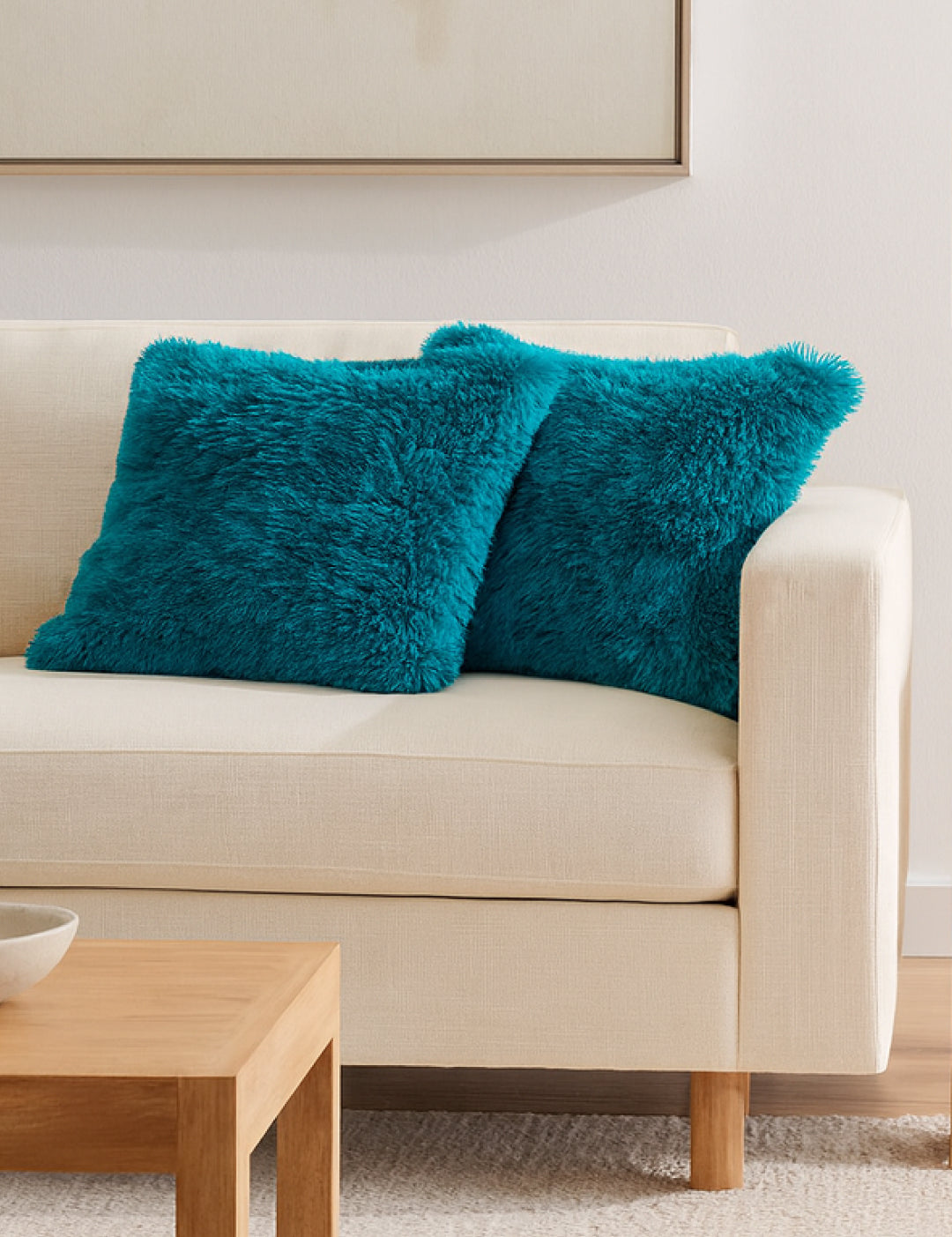 A beige sofa with two Juego De Cojín Everest by Intima Hogar in turquoise sits beside a light wood coffee table in a modern, minimal living room. Above the sofa, a large framed artwork is partially visible on the wall.