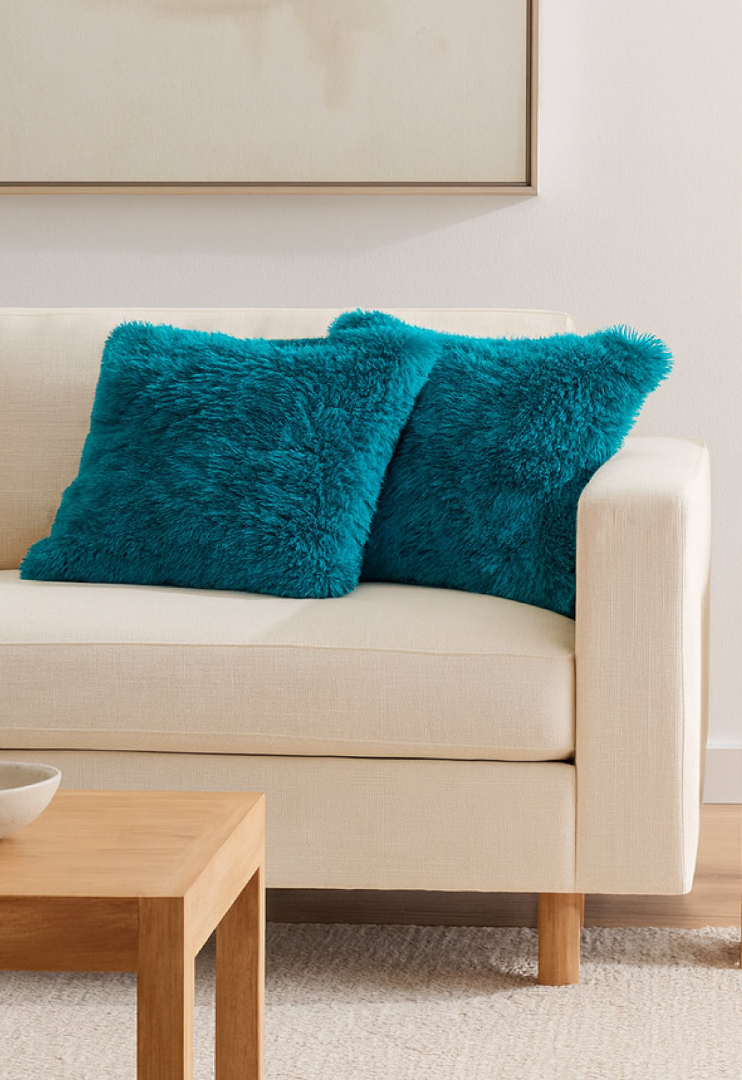 A beige sofa with two Juego De Cojín Everest by Intima Hogar in turquoise sits beside a light wood coffee table in a modern, minimal living room. Above the sofa, a large framed artwork is partially visible on the wall.