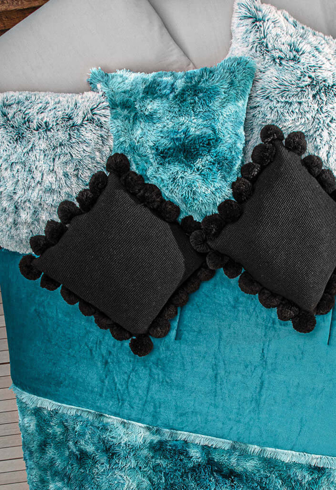 A bed topped with the Juego De Cojín Everest by Intima Hogar, featuring a teal velvet and faux fur blanket, turquoise cushions, and two black textured pillows with pom-pom trim, all neatly arranged.