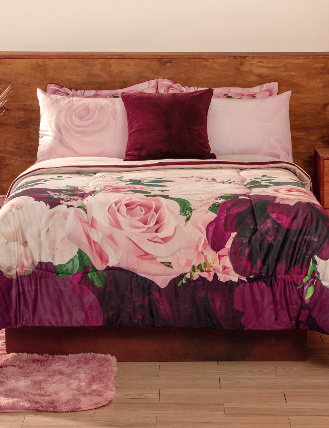 A neatly made bed with the Coordinado de Cobertor Supremo Granate by Intima Hogar, matching pink pillowcases, a dark purple accent pillow, wooden headboard, light wood floor, and a pink rug.