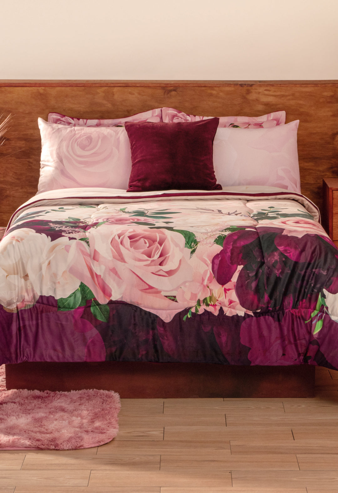 A neatly made bed with the Coordinado de Cobertor Supremo Granate by Intima Hogar, matching pink pillowcases, a dark purple accent pillow, wooden headboard, light wood floor, and a pink rug.
