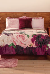 A neatly made bed with the Coordinado de Cobertor Supremo Granate by Intima Hogar, matching pink pillowcases, a dark purple accent pillow, wooden headboard, light wood floor, and a pink rug.