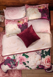 A neatly made bed features the Intima Hogar Coordinado de Cobertor Supremo Granate with floral bedding in pink, white, and purple tones (flores tonos rosa), four matching pillows, a solid burgundy pillow, wooden headboard, and nightstand.