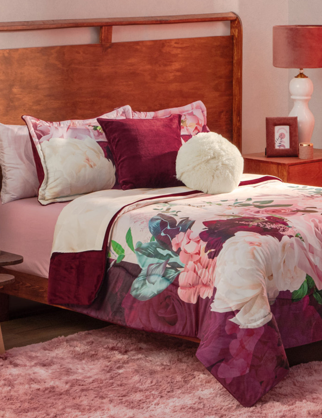A cozy bed with the Coordinado de Cobertor Supremo Granate by Intima Hogar, white and burgundy pillows, a fuzzy round cushion, and a wooden headboard. The room has a pink rug, red nightstand with lamp, and soft pink walls.