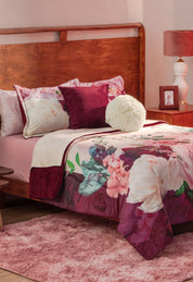 A cozy bed with the Coordinado de Cobertor Supremo Granate by Intima Hogar, white and burgundy pillows, a fuzzy round cushion, and a wooden headboard. The room has a pink rug, red nightstand with lamp, and soft pink walls.