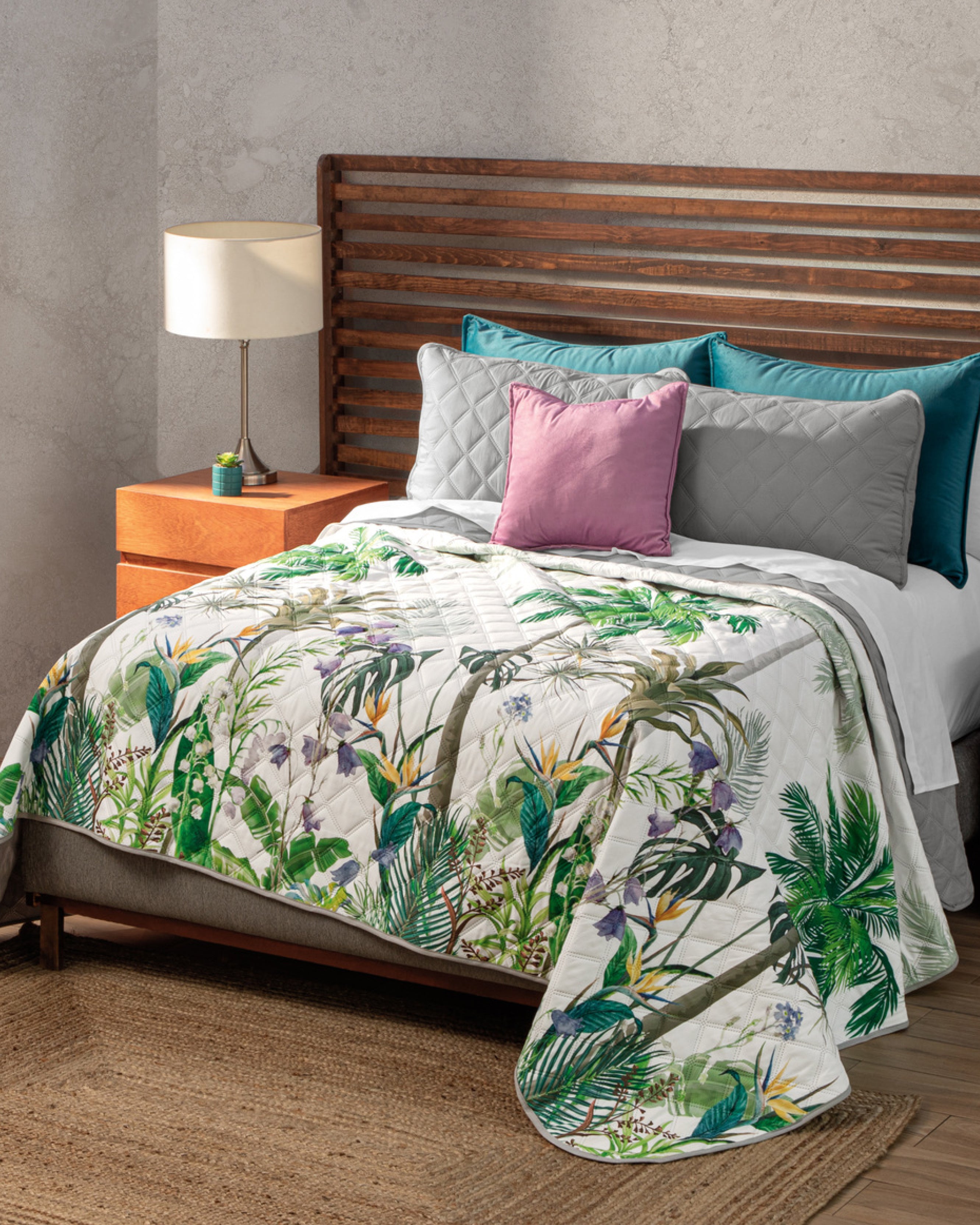 A neatly made bed with gray, teal, and lavender pillows, the Intima USA Juego de Edredón Tropical featuring green leaves and flowers, a wooden headboard, a wooden nightstand with a lamp, and a woven rug on the floor.