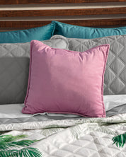 A pink square pillow rests on a bed with gray textura ultra suave bedding, teal pillows by a wooden headboard, and the Intima USA Juego de Edredón Tropical—a white comforter with green leaf prints.