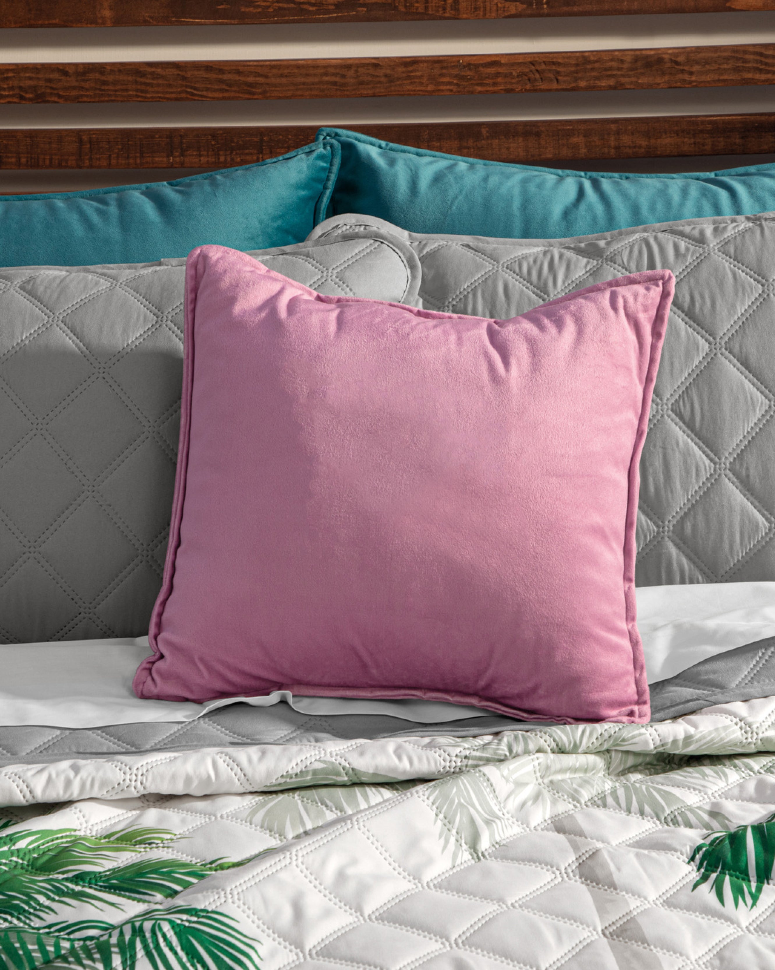 A pink square pillow rests on a bed with gray textura ultra suave bedding, teal pillows by a wooden headboard, and the Intima USA Juego de Edredón Tropical—a white comforter with green leaf prints.