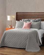 A neatly made bed features the Juego de Edredón Tropical by Intima USA in gray with textura ultra suave, fluffy gray pillows, and a pink accent pillow. A wooden headboard, nightstand with lamp, flowers, and teacup complete the cozy setting.