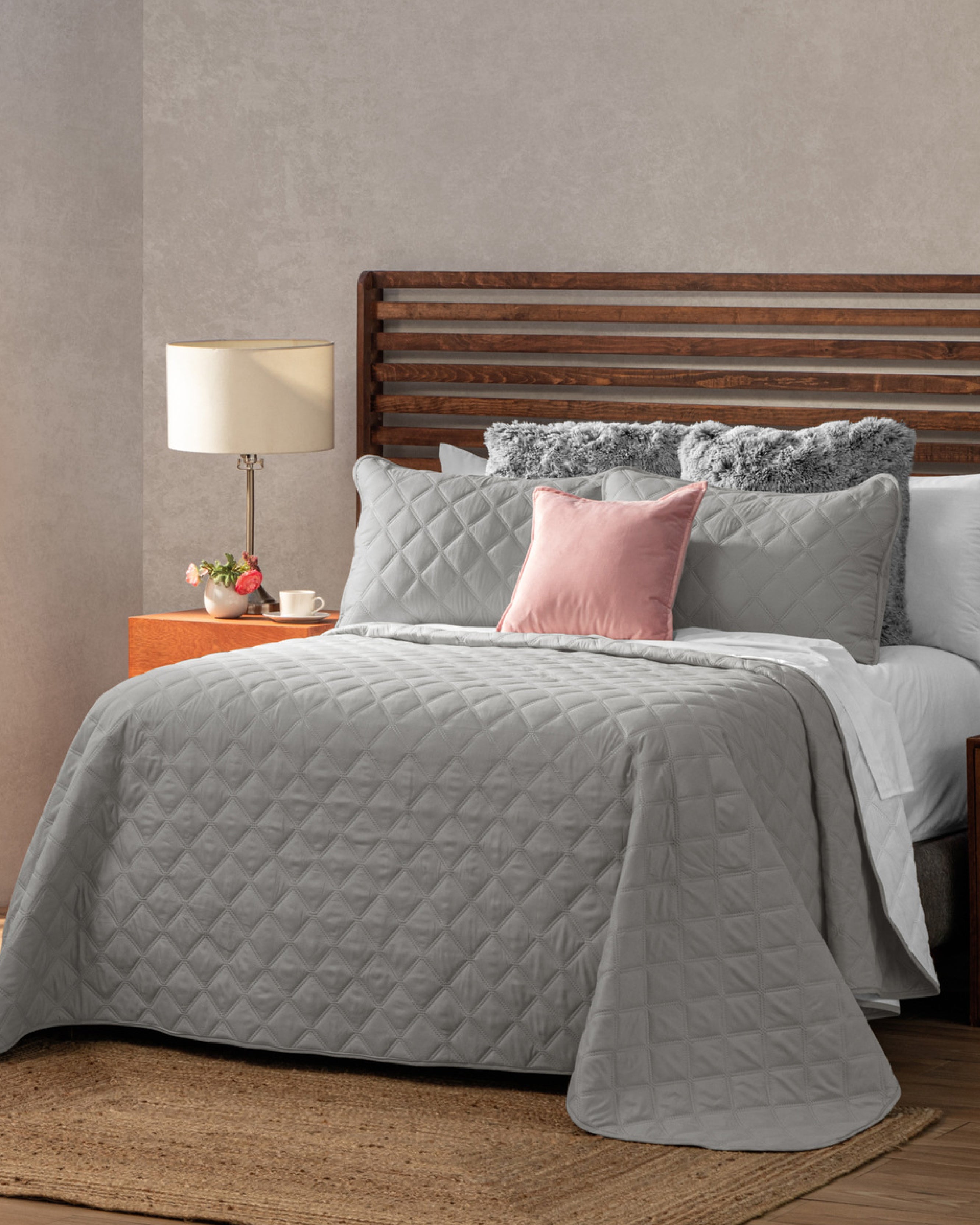 A neatly made bed features the Juego de Edredón Tropical by Intima USA in gray with textura ultra suave, fluffy gray pillows, and a pink accent pillow. A wooden headboard, nightstand with lamp, flowers, and teacup complete the cozy setting.