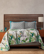 A neatly made bed with a wooden slatted headboard, two gray quilted pillows, and the Juego de Edredón Tropical by Intima USA featuring ultra-soft texture. A wooden nightstand with lamp, book, and turquoise glass stands to the right.