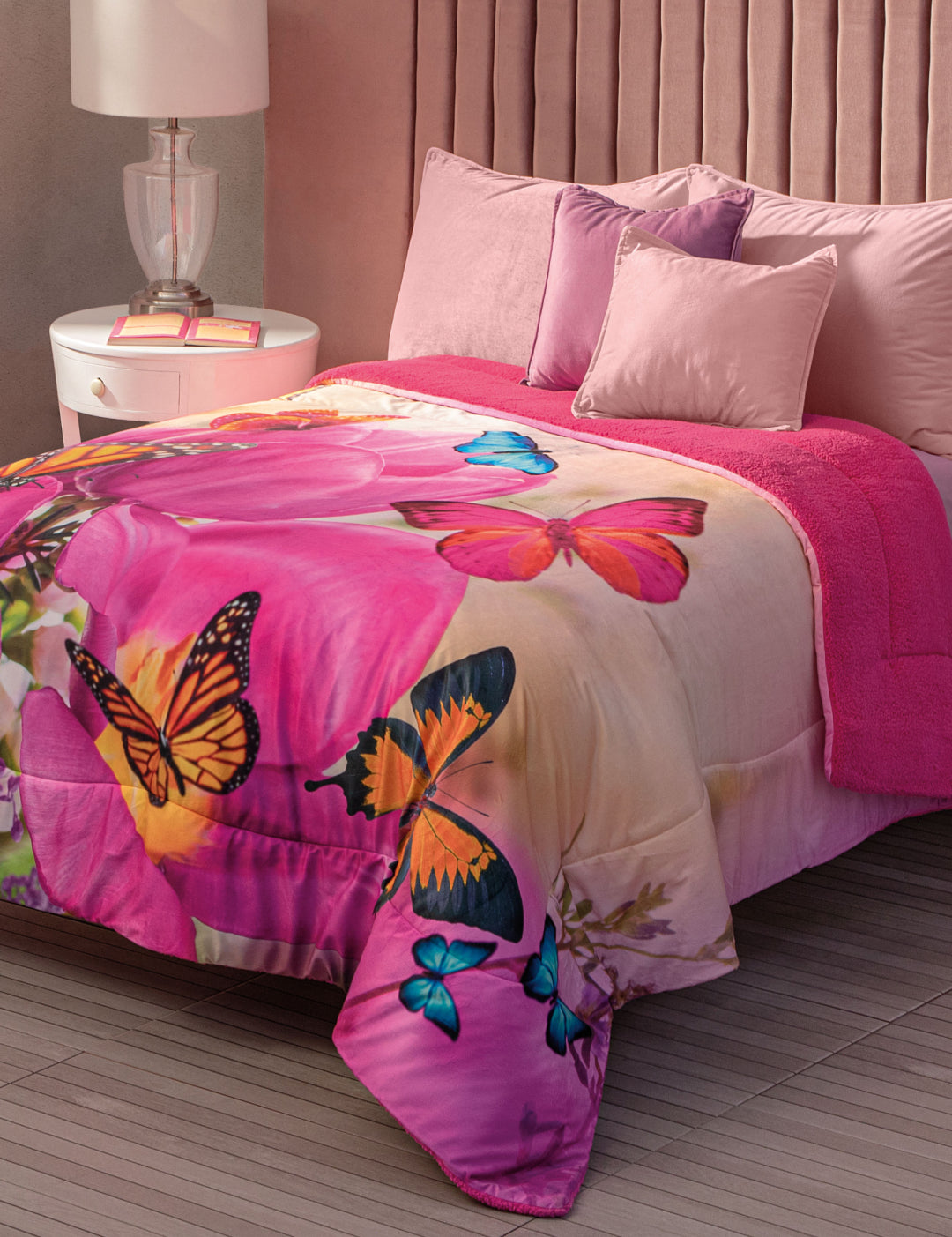 A neatly made bed with Intima Hogar’s Cobertor Digital Ultra Suave Mariposa-Desc, pink pillows, a velvet headboard, a bedside table with a lamp, and a soft pink franela sherpa blanket.