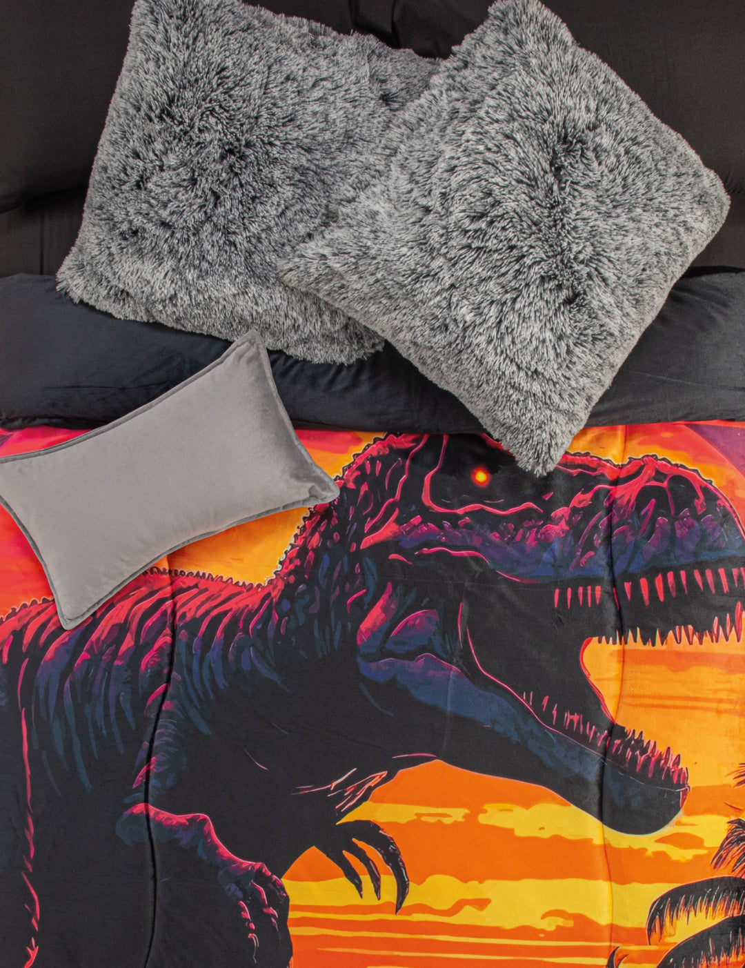 A bed with black sheets, two large gray fluffy pillows, a small gray pillow, and the Cobertor Digital Suave Dinosaurio Rex by Intima Hogar featuring roaring dinosaurs at sunset with palm trees and a vibrant prehistoric design.