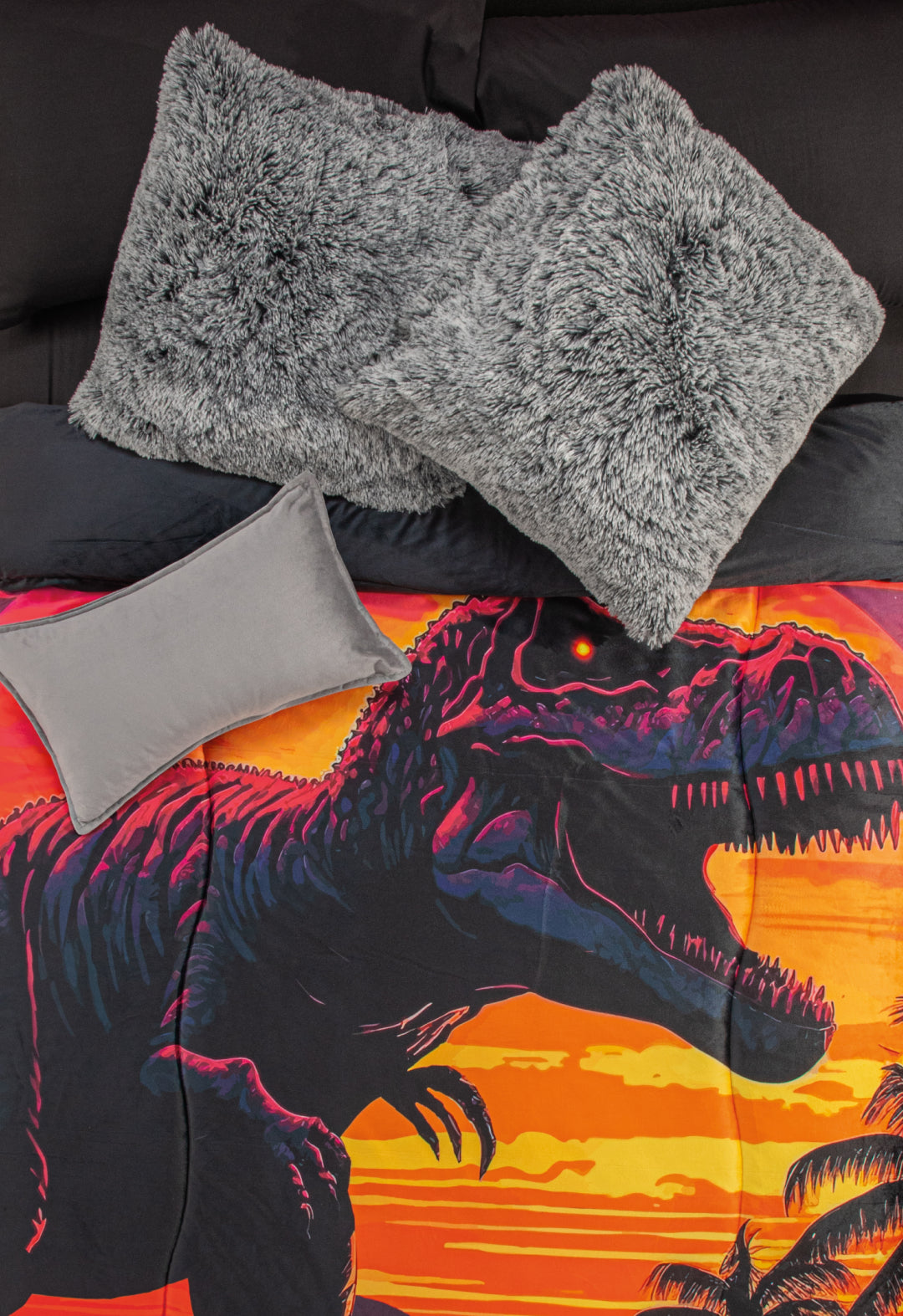 A bed with black sheets, two large gray fluffy pillows, a small gray pillow, and the Cobertor Digital Suave Dinosaurio Rex by Intima Hogar featuring roaring dinosaurs at sunset with palm trees and a vibrant prehistoric design.