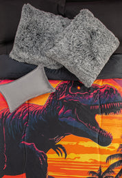 A bed with black sheets, two large gray fluffy pillows, a small gray pillow, and the Cobertor Digital Suave Dinosaurio Rex by Intima Hogar featuring roaring dinosaurs at sunset with palm trees and a vibrant prehistoric design.