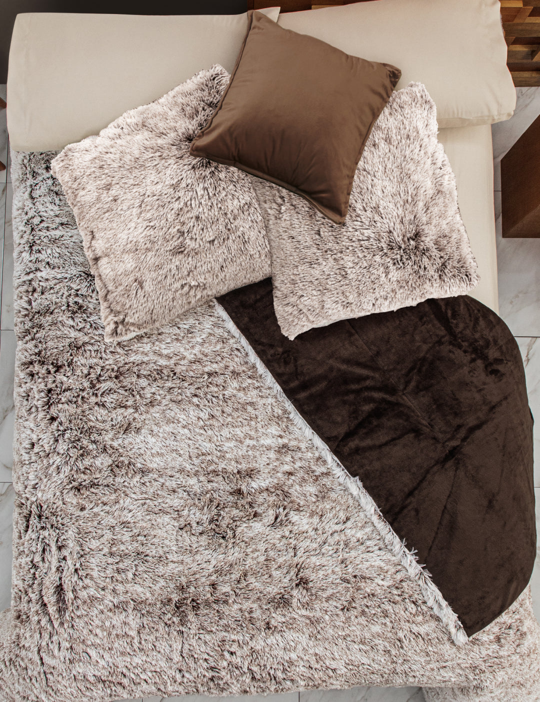 A neatly made bed features the Intima Hogar Juego de Cojines de Lujo Glacial, with plush beige pillows, a brown satin decorative cushion, and a cozy beige and brown blanket, all set atop a marble floor.