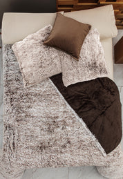 A neatly made bed features the Intima Hogar Juego de Cojines de Lujo Glacial, with plush beige pillows, a brown satin decorative cushion, and a cozy beige and brown blanket, all set atop a marble floor.