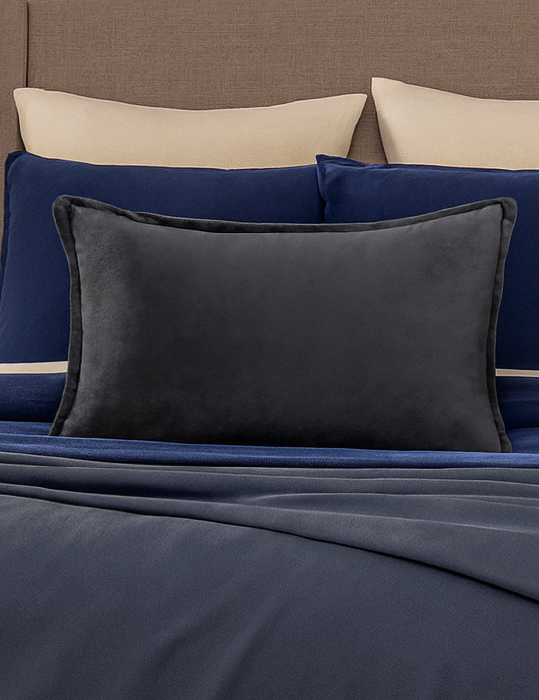 A neatly made bed with beige and navy blue pillows, a large Funda de Cojín Rectangular Sueños Gris Oxford by Intima Hogar in front, and a navy blue blanket. The beige headboard and this cozy funda add extra warmth to the room.