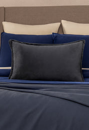 A neatly made bed with beige and navy blue pillows, a large Funda de Cojín Rectangular Sueños Gris Oxford by Intima Hogar in front, and a navy blue blanket. The beige headboard and this cozy funda add extra warmth to the room.